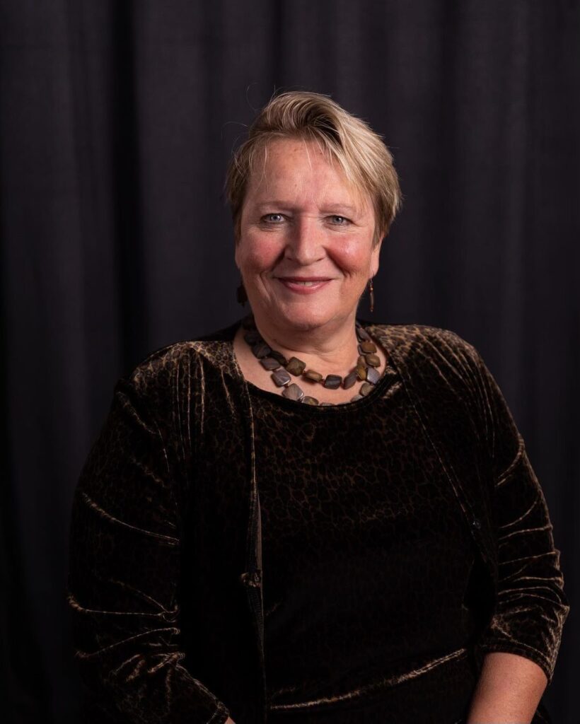 A white woman with blond hair is smiling at the camera. She is wearing a dark shirt and a necklace. 